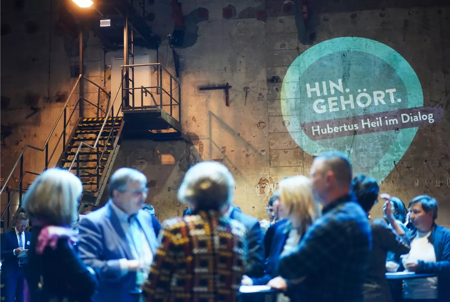 Foto von einer Veranstaltung. Im Vordergrund diskutieren Menschen an einem Stehtisch. Im Hintergrund ist das Logo "Hin. gehört" an die Wand projiziert.